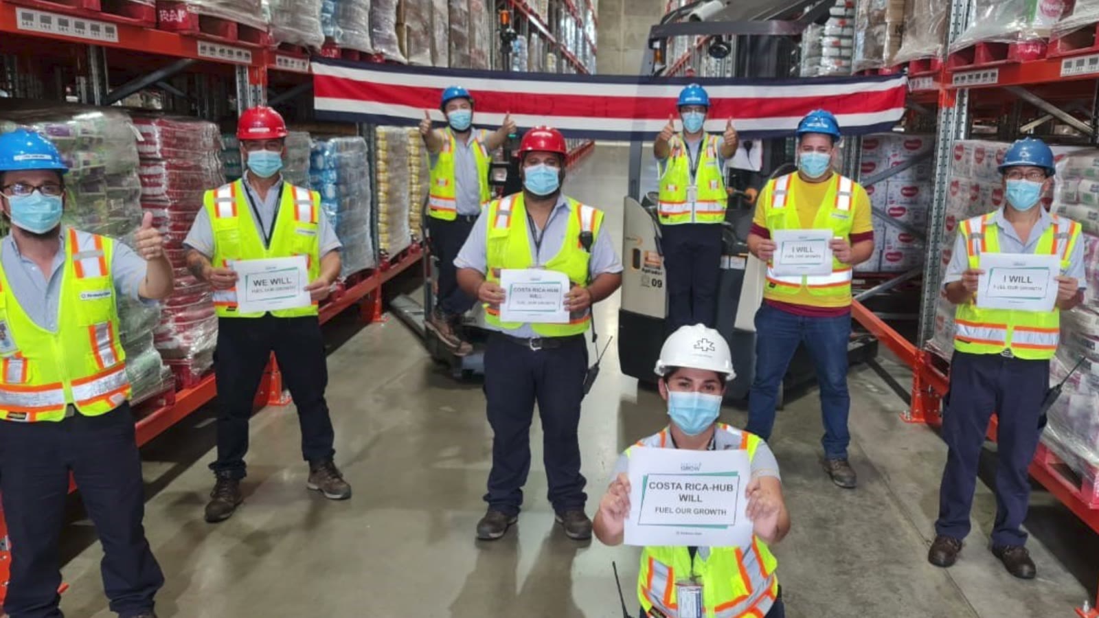 A team in hard hats and safety vests holding signs in a warehouse aisle